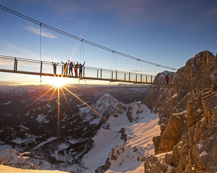 A group of excited hikers poses on a suspended bridge in Schladming-Dachstein, framed by a stunning mountain sunset.