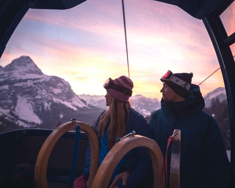 A couple with 2 sledges look out of the gondola lift at the mountains at sunset.