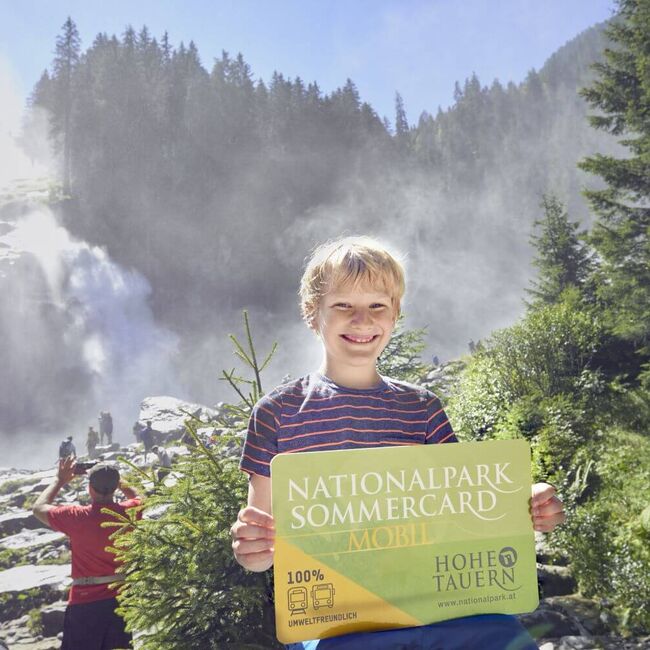 Junge stands in front of the Krimml Waterfalls with the National Park Summer Card in his hand.