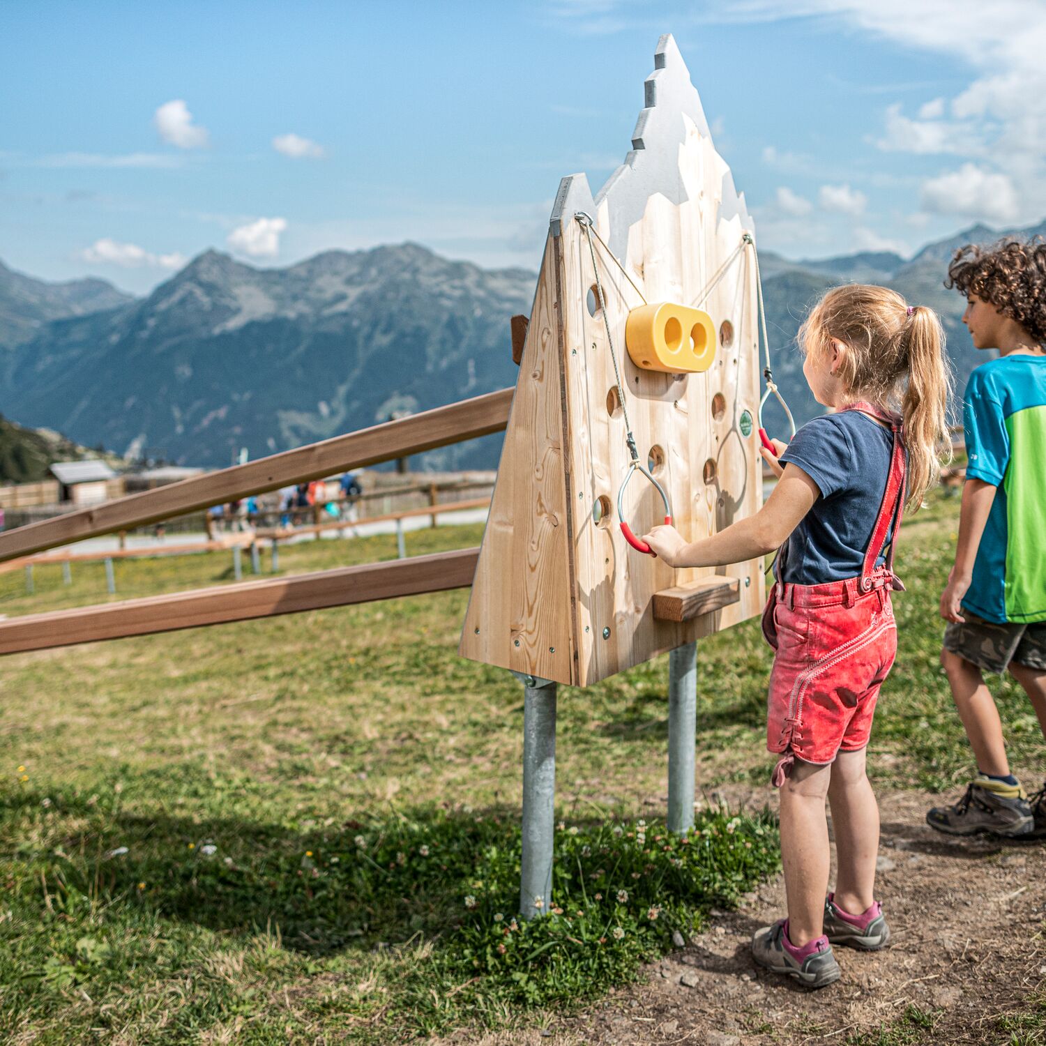 Twee kinderen in wandelkleding spelen met een houten knikkerbaan op een alpenweide, met imposante bergen op de achtergrond
