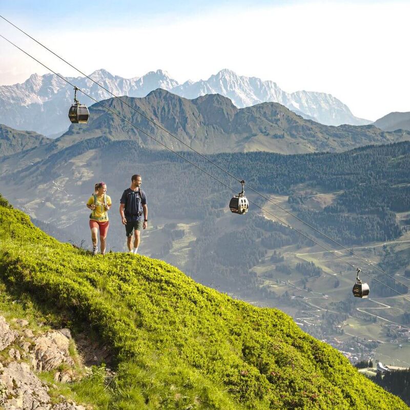 Über 2 Wanderern fährt die Sommerbergbahn, im Hintergrund 2 imposante Bergkämme.