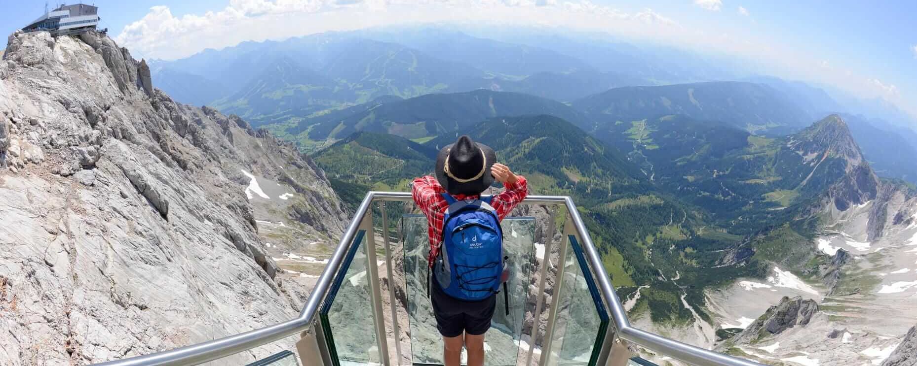A hiker stands on a glass platform, gazing at breathtaking mountain views from Dachstein's peak.