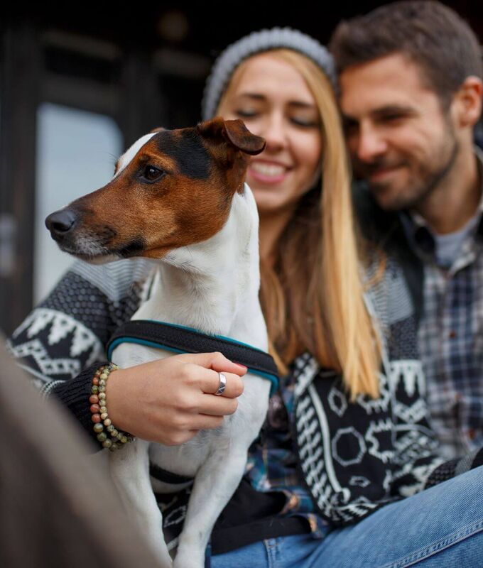 Pärchen im Winterurlaub sitzt auf der Terrasse, hält den Hund im Schoß und genießt ein wärmendes Heißgetränk