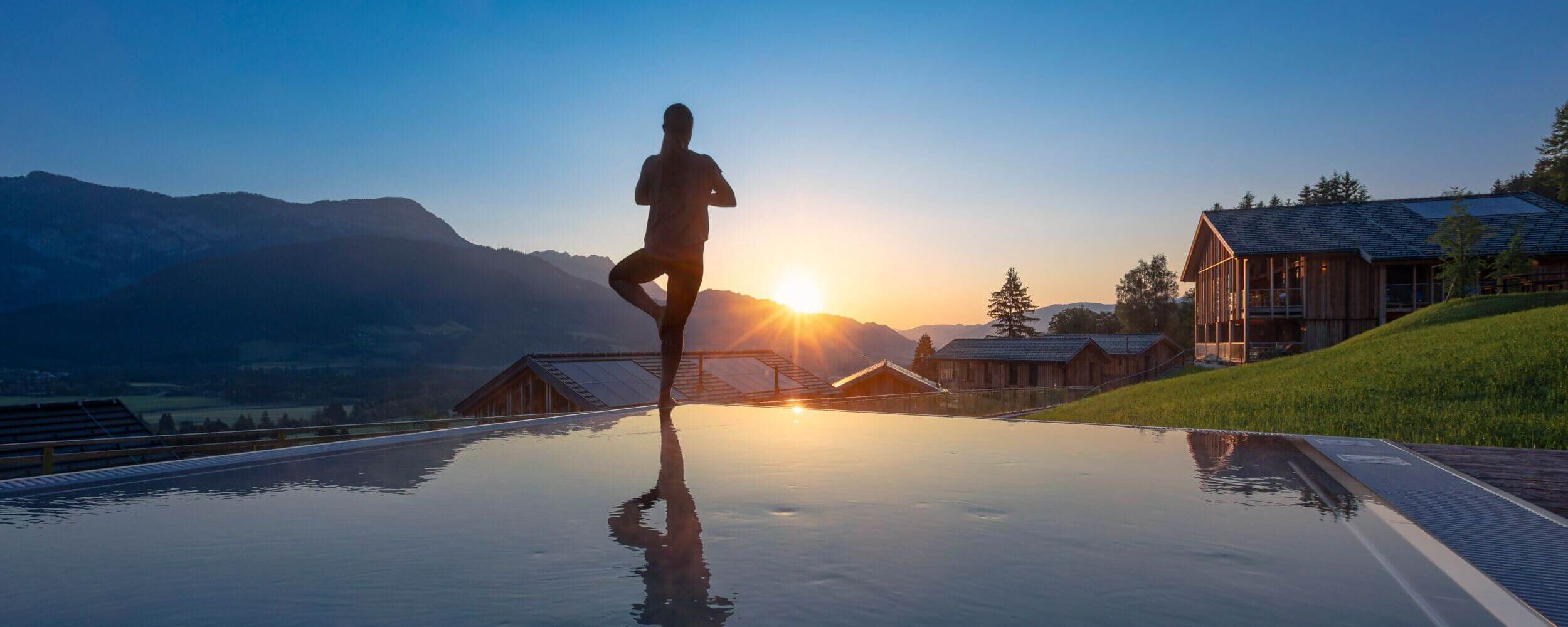 A person practices yoga at sunrise by a tranquil pool with mountains and cabins in the background.