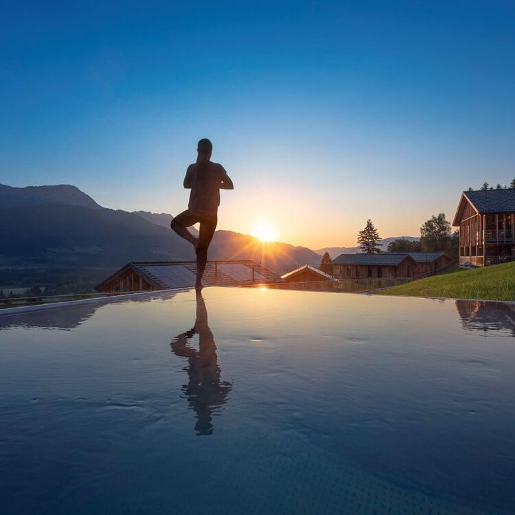 Frau beim Yoga am privaten Pool ihres Ferienhauses, im Hintergrund Berge und Sonnenaufgang.