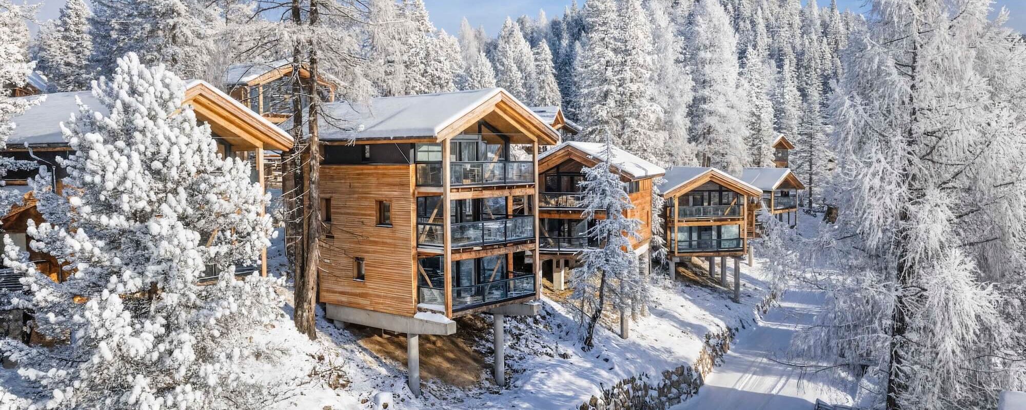 Winterliche Außenansicht der Naturchalets Turracher Höhe mit verschneitem Chalet inmitten des Waldes und tief verschneiter alpiner Umgebung.