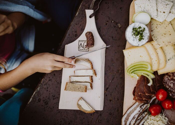 Brettljause mit frischem Bauernbrot, verschiedenen Käsesorten, Speck, Wurst, Tomaten, Äpfeln und Schnittlauchaufstrich, serviert im Salzburger Almsommer