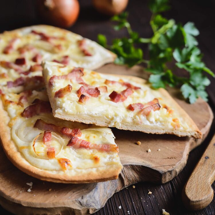 Drei köstliche Flammkuchen mit Speck und Zwiebeln auf einem Holzbrett, umgeben von frischen Kräutern.