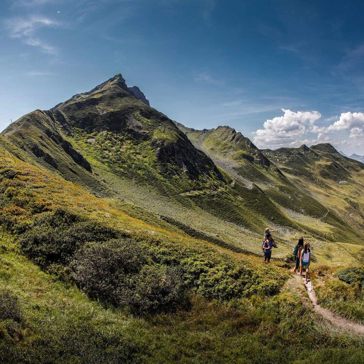 Hikers traverse a scenic trail in the Spieljoch mountains, surrounded by vibrant greenery and stunning alpine views.