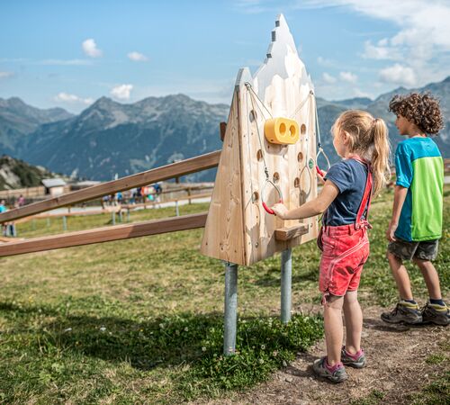 Zwei Kinder in Wanderkleidung spielen auf einer Alm bei einer Kugelbahn aus Holz, im Hintergrund ragen imposante Berge in den Himmel