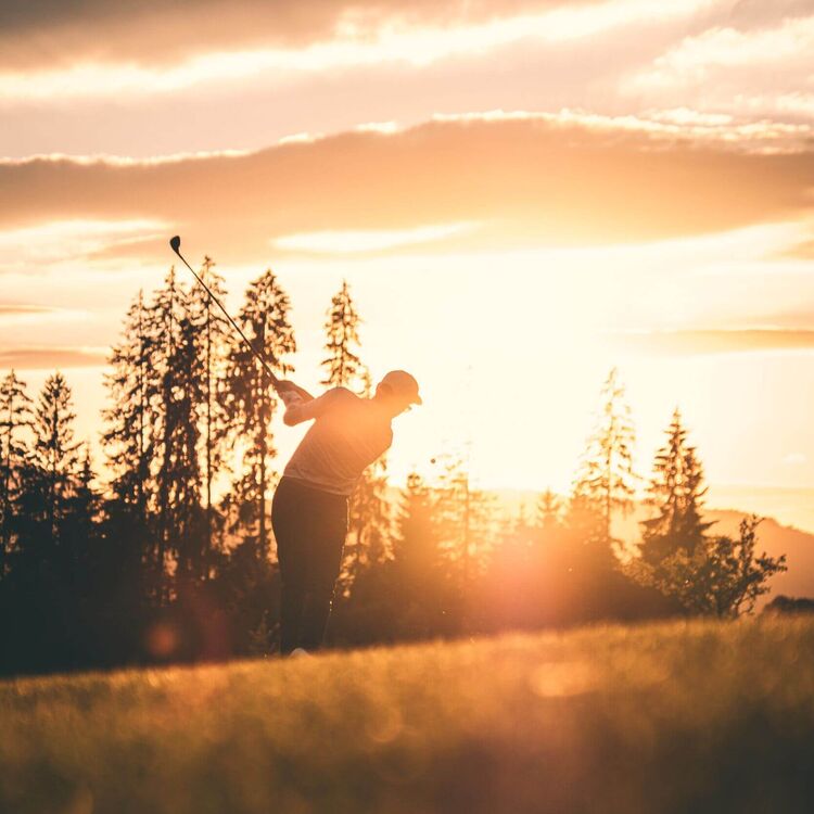 A golfer swings against a stunning sunset, silhouetted by trees and bathed in warm, golden light.