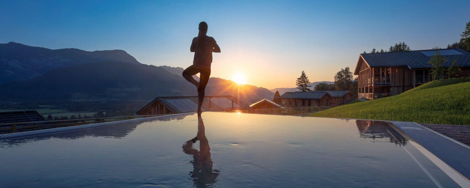 Frau beim Yoga am privaten Pool ihres Ferienhauses, im Hintergrund Berge und Sonnenaufgang.