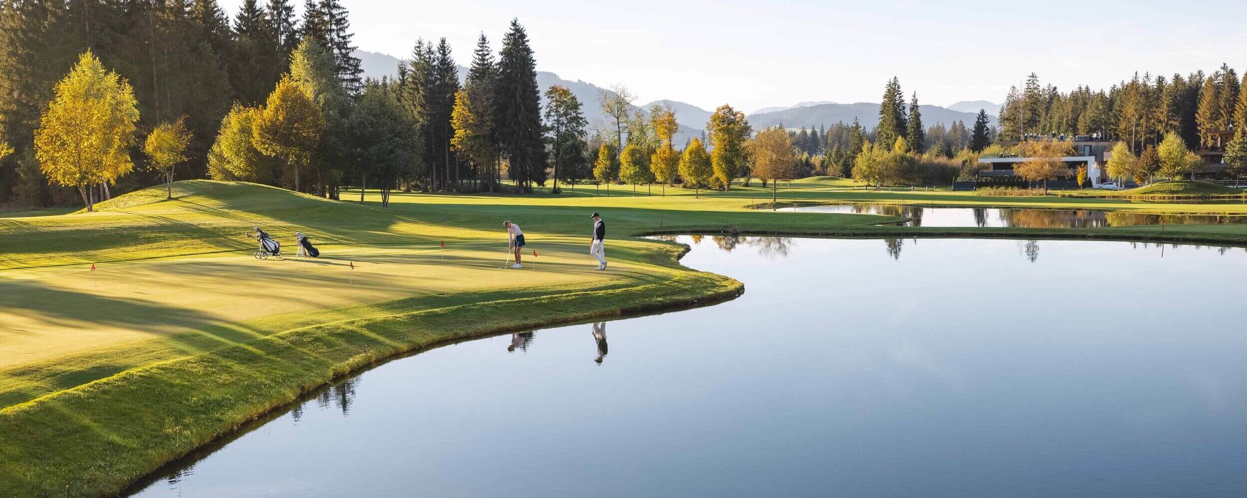 Two golfers practice their putting on a tranquil golf course, reflecting autumn colors in the calm water nearby.