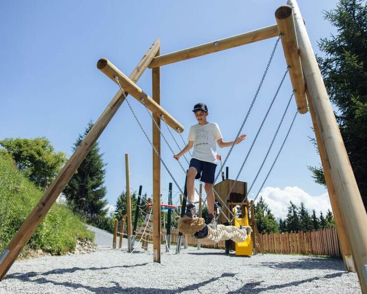 A child balances on a rope bridge at Schmidolins' playground, surrounded by trees and a clear blue sky.