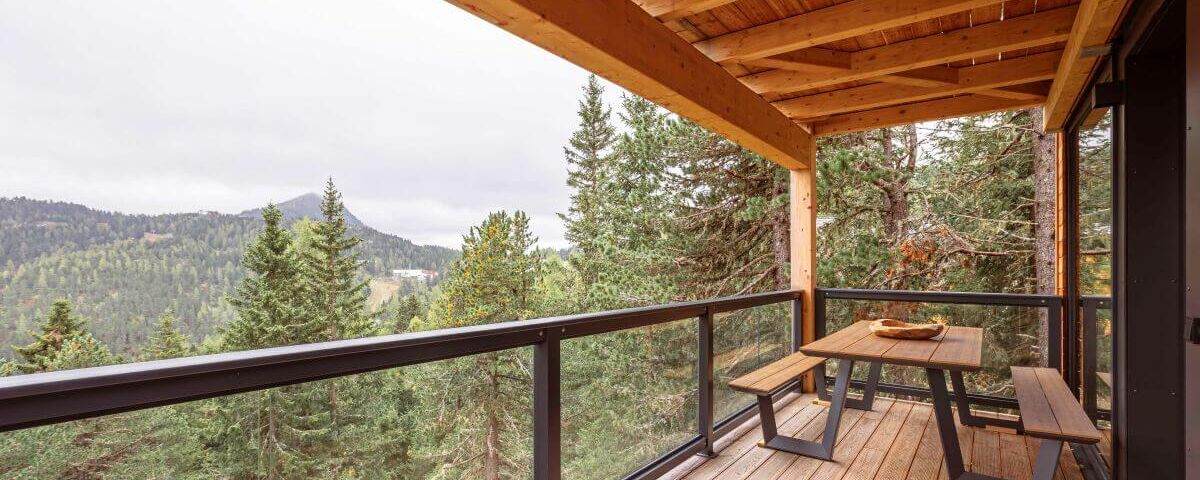 A cozy wooden deck with a table offers stunning views of lush green forests and distant mountains at a mountain resort.