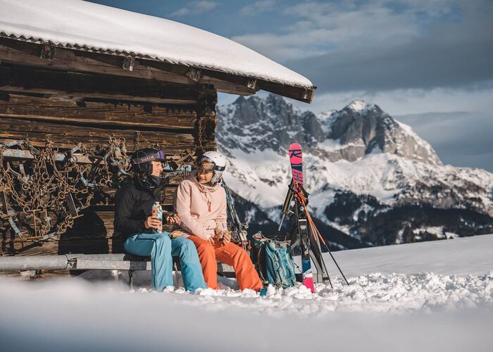 Zwei Skifahrerinnen entspannen vor einer Berghütte am Wilden Kaiser und genießen die winterliche Aussicht.