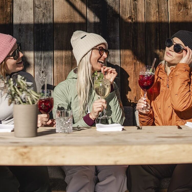3 friends in ski outfits sit in the sun in front of a ski hut.