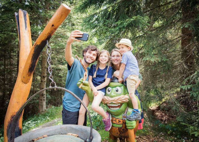 A family takes a selfie with a friendly green statue in the scenic Tiroler Zugspitze forest.