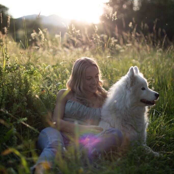 Claudia sitzt im grünen Feld und lächelt glücklich neben ihrem Samojedenhund.