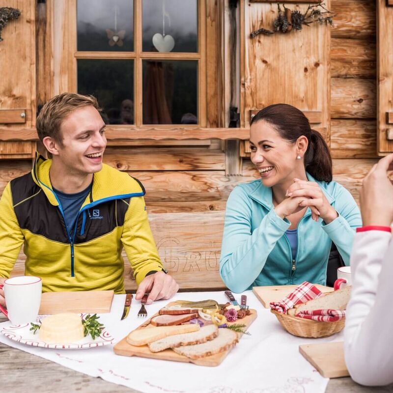 Three people sit at a table in a Tyrolean mountain pasture and enjoy regional products such as bread and bacon