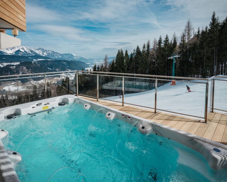 A luxurious outdoor jacuzzi overlooks snowy slopes and mountain scenery, with a skier enjoying the winter landscape nearby.