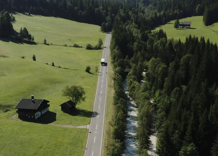 Bus travelling on a country road surrounded by green meadows, forest and river.