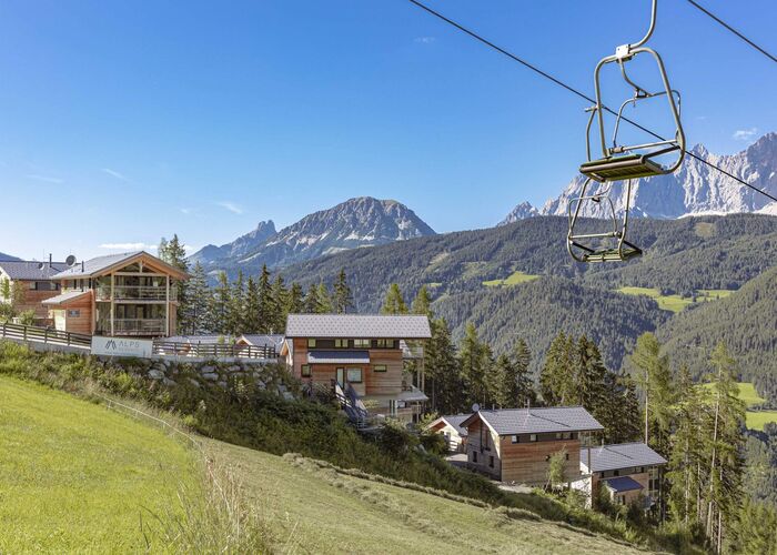 Ein Sessellift schwebt über malerische Chalets in den Alpen, umgeben von grünen Wiesen und majestätischen Bergen.