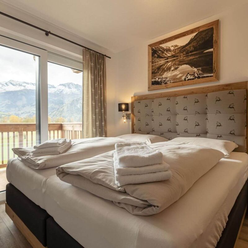 Licht slaapkamer met comfortabel boxspring tweepersoonsbed, houtelementen, gevouwen handdoeken op het bed, gestoffeerd hoofdbord, nachtkastjes en een balkon met uitzicht op herfstlandschap en besneeuwde bergen