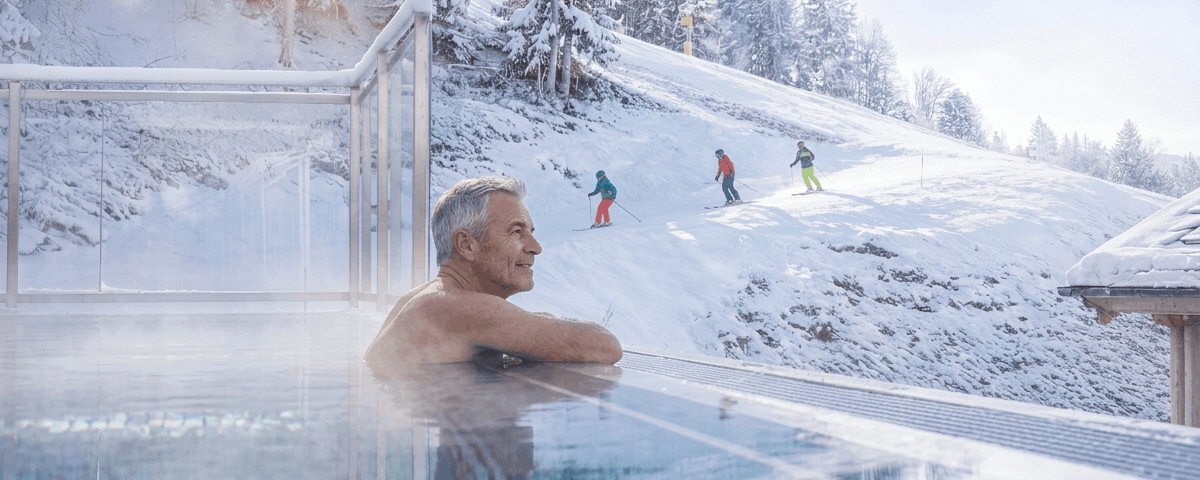 A relaxed older man enjoys a warm pool view while skiers descend snowy slopes in a winter landscape.