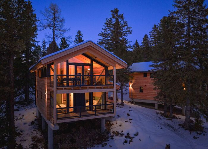 Außenansicht eines verschneiten Chalets der Naturchalets Turracher Höhe by ALPS RESORTS am Abend, stimmungsvoll beleuchtet und umgeben von winterlichem Wald.
