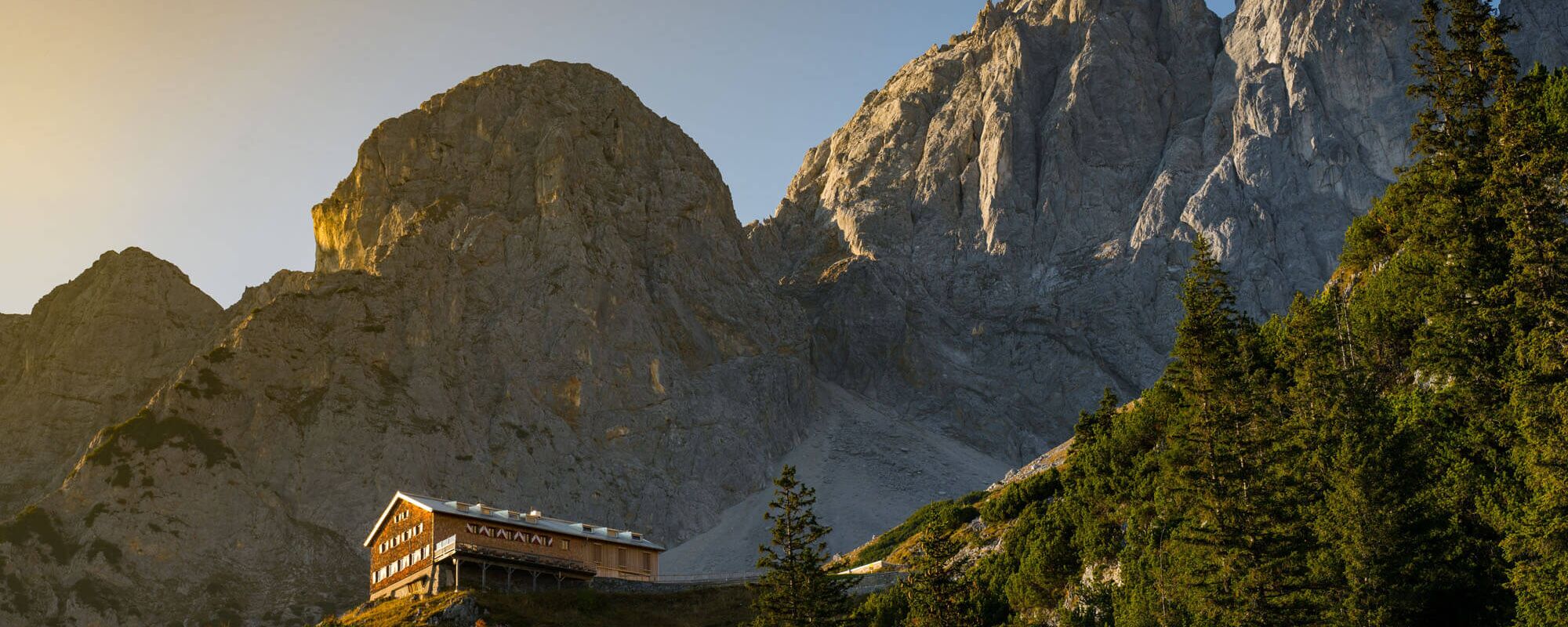 Die Gruttenhütte schimmert im Sonnenlicht, umgeben von majestätischen Bergen des Wilden Kaisers und dichten Wäldern.