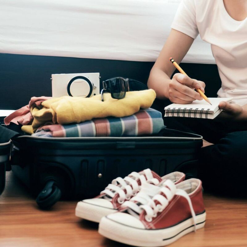 Young woman with a checklist sits on the floor in front of her open, fully packed suitcase.