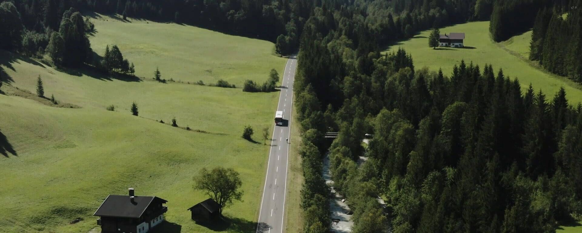 Bus fährt auf einer Landstraße umgeben von grünen Wiesen, Wald und Fluss.