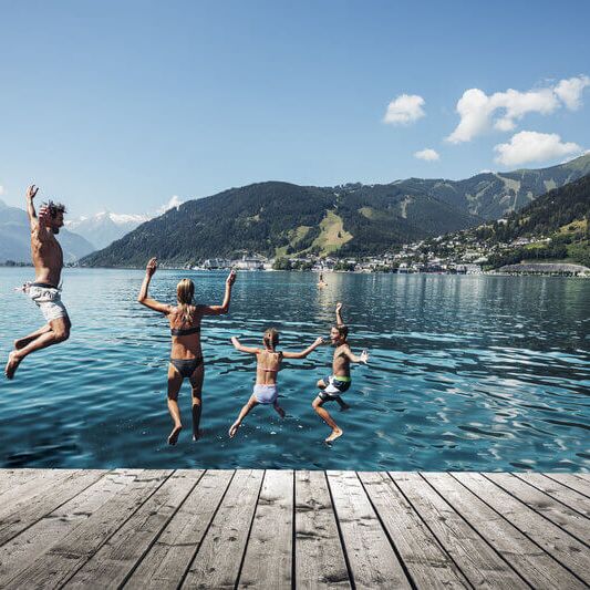 Familie springt in het meer van Zell, met de Kitzsteinhorn en de stad Zell am See op de achtergrond.