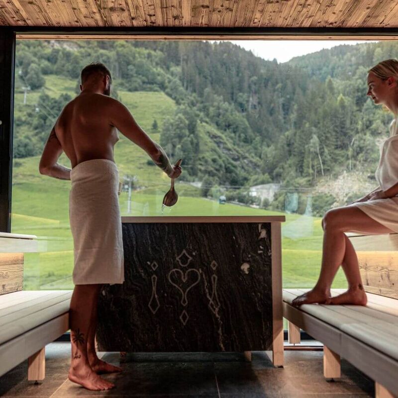 Pärchen beim Aufguss in einer Panoramasauna mit Blick auf die Bergbahn