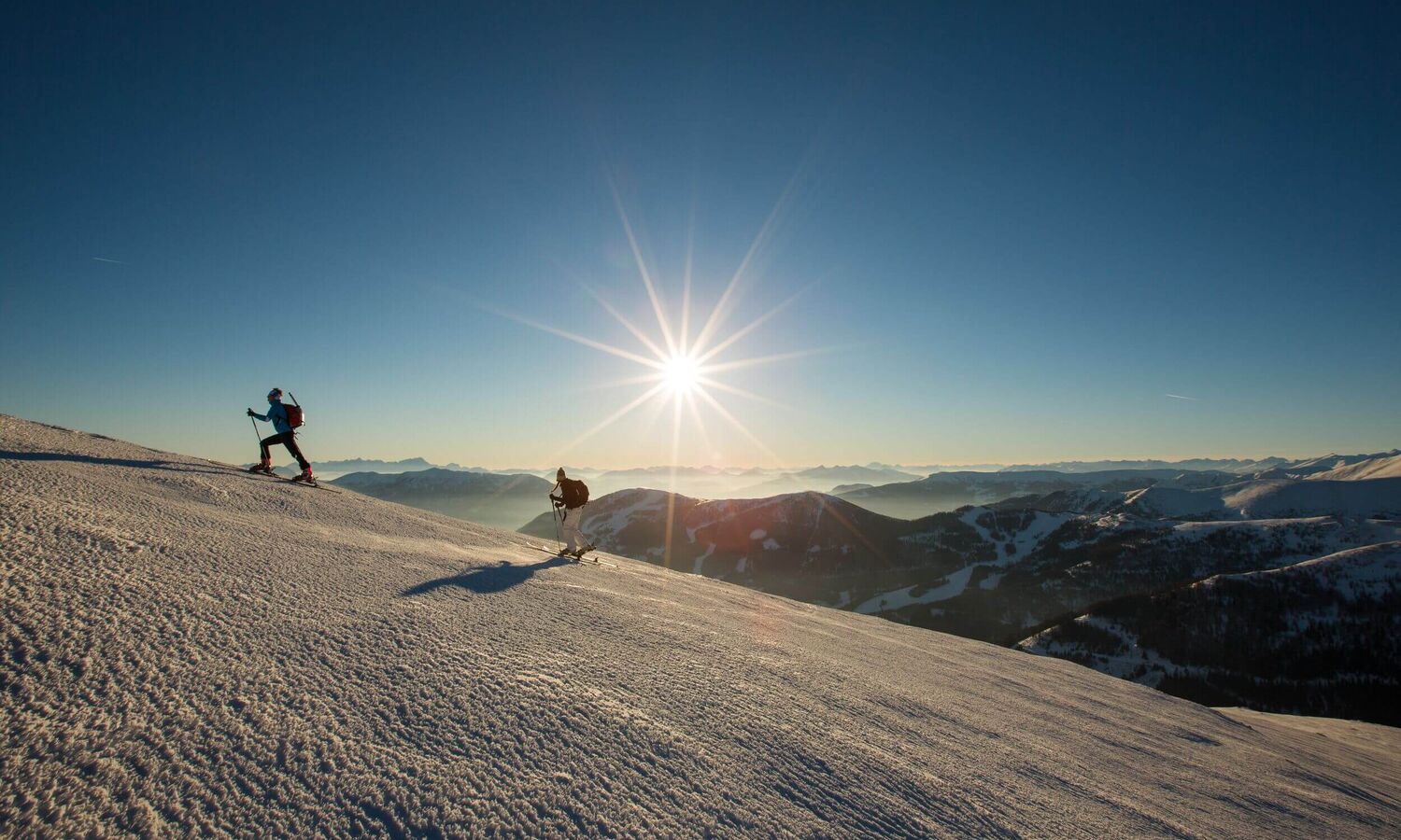 Zwei Skitourengeher steigen bei Sonnenaufgang durch die verschneiten Nockberge