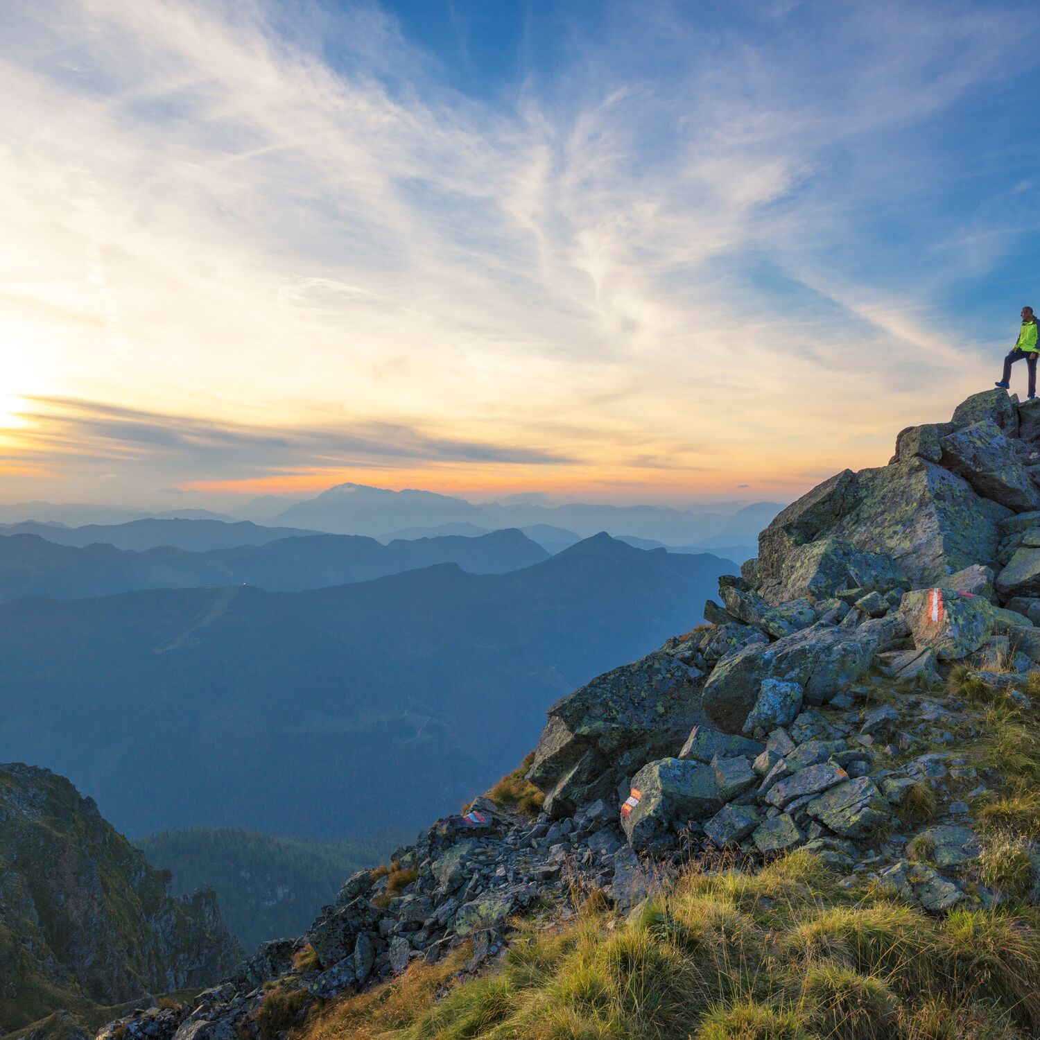 Twee wandelaars staan bij een bergtopkruis en kijken uit over het avondlandschap met weids panorama en warm licht