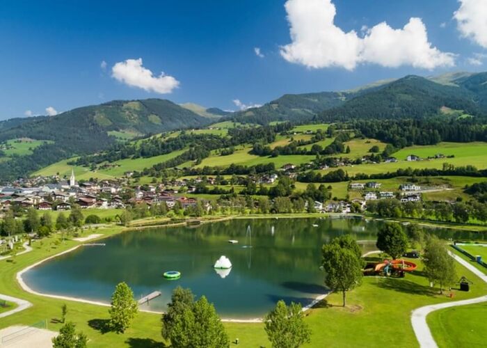 Sommerliche Aussicht auf einen Badesee im Familiendorf, umgeben von Bergen und einer malerischen Landschaft.