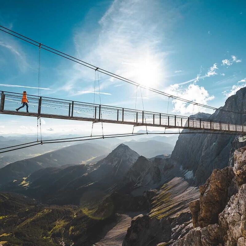 A person walks across a stunning suspension bridge, surrounded by majestic mountains and a bright blue sky.