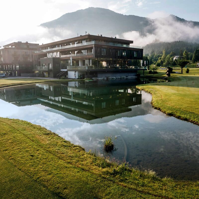 Mountain resort reflects in a tranquil pond, surrounded by lush greenery and misty mountains.