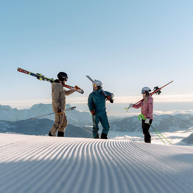 3 skier friends stand in the sun on the fresh piste above the sea of fog.