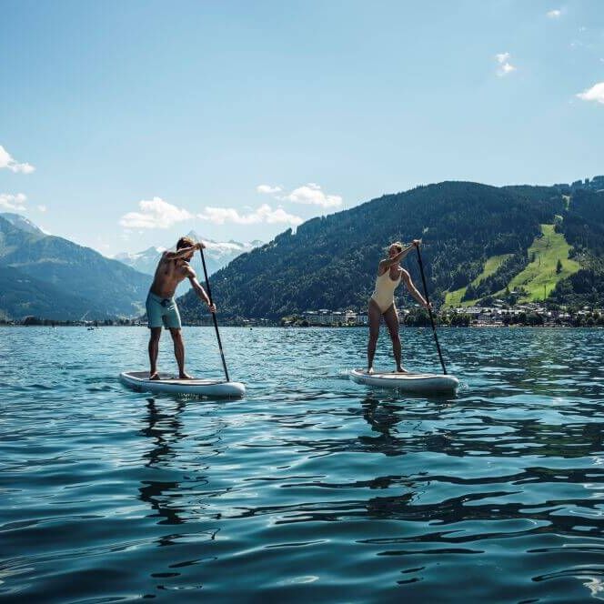 Two people paddleboard on a serene lake surrounded by lush mountains under a bright blue sky.