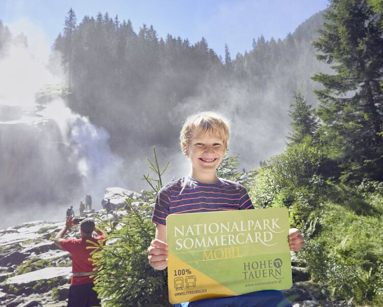 Junge stands in front of the Krimml Waterfalls with the National Park Summer Card in his hand.