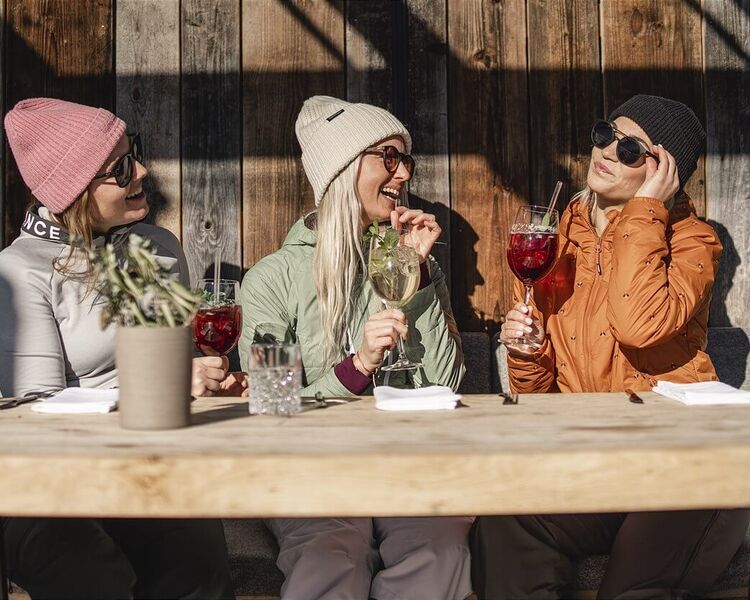3 friends in ski outfits sit in the sun in front of a ski hut.
