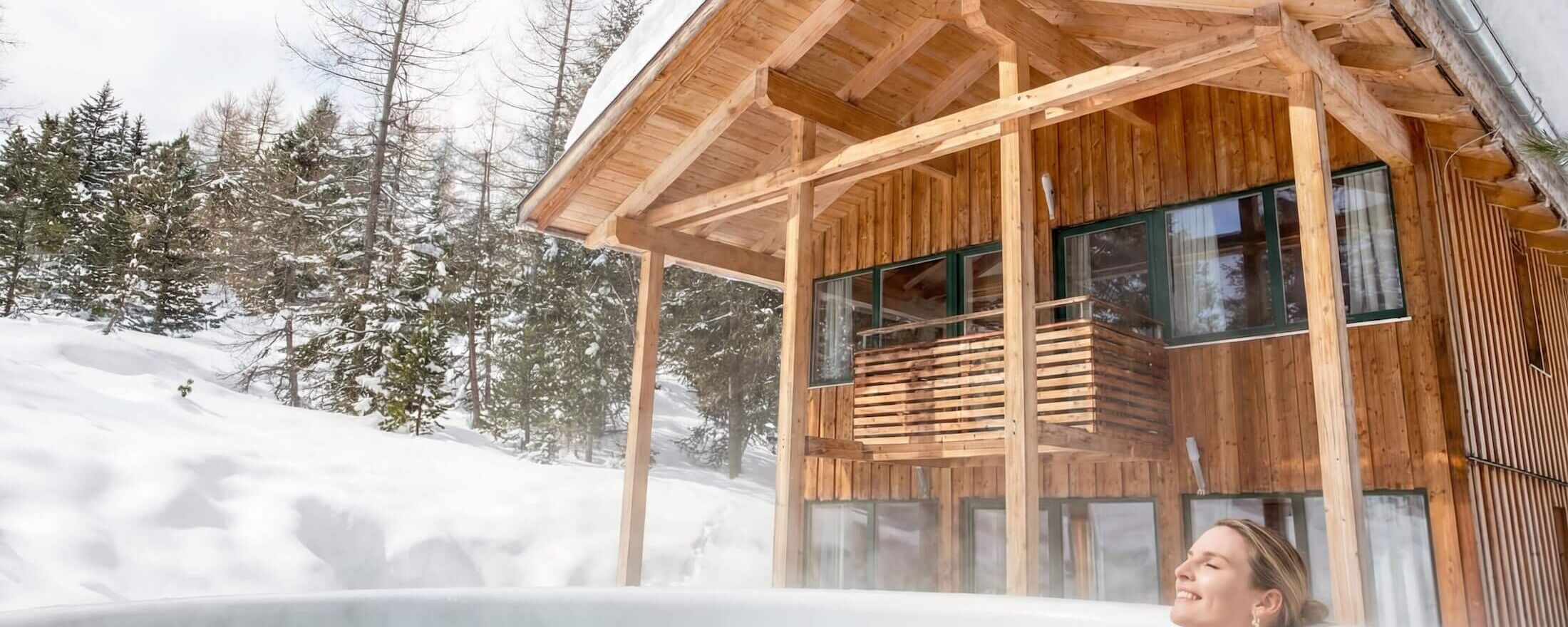 A woman relaxes in a hot tub outside a cozy wooden chalet amid snowy Alps scenery.