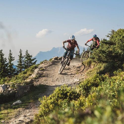Two downhill bikers riding dynamically down an alpine trail, surrounded by impressive mountain scenery