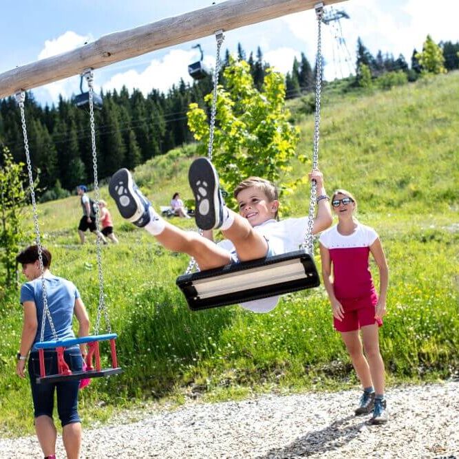 Children enjoy swinging at the Maiskogel playground, with lush greenery and mountains in the background. | © Kitzsteinhorn