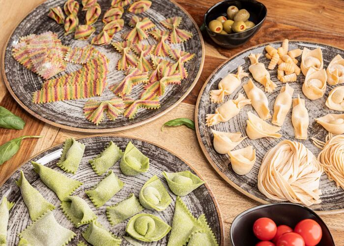 Bunte Pasta-Sorten auf Tellern, darunter gefüllte Teigtaschen und frische Nudeln, dekoriert mit Oliven und Tomaten.