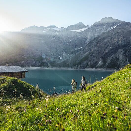 Twee wandelaars lopen langs een rustieke berghut over een bloeiende alpenweide met uitzicht op een turquoise stuwmeer en het omliggende berglandschap in Salzburger Land.