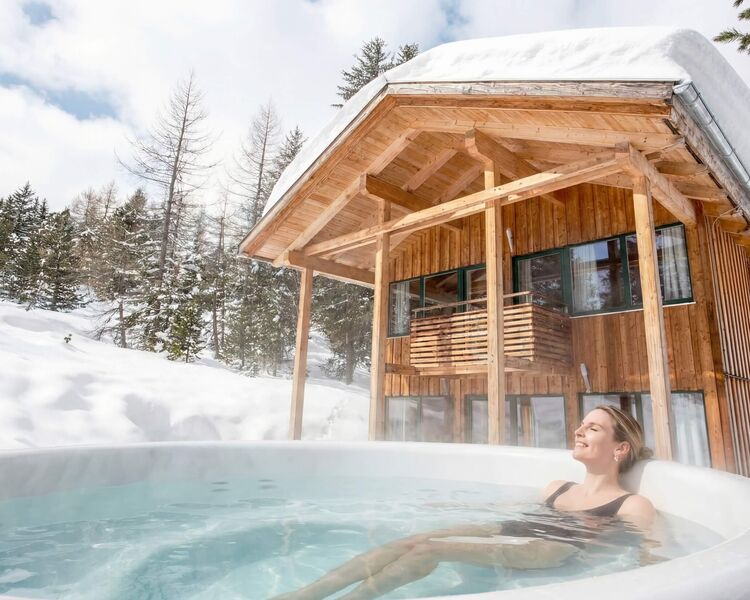 Eine Frau entspannt in einem Whirlpool vor einem Holzhaus in einer schneebedeckten Alpenlandschaft.
